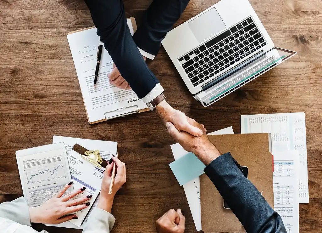 Dos personas se dan la mano sobre una mesa con documentos, gráficas y una computadora portátil